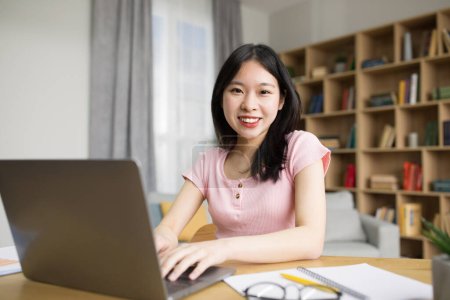 Téléchargez les photos : Concept technologique. Femme asiatique excitée assis au bureau et regardant la caméra, en utilisant un ordinateur portable dans le salon de son appartement moderne. Bonne femme coréenne posant pour la photo à la maison - en image libre de droit