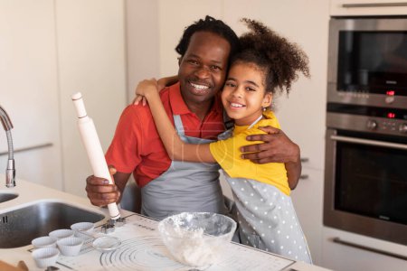Foto de Retrato de Happy Black Girl y su padre abrazándose en el interior de la cocina, alegre papá afroamericano y su hija usando delantales posando en la cámara mientras hornean juntos en casa, espacio libre - Imagen libre de derechos