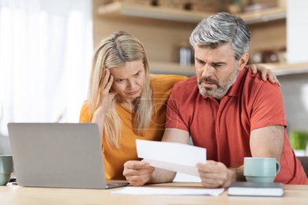 Photo for Anxious middle aged husband and wife sitting at table in front of computer, checking correspondence, upset with their debts, kitchen interior, copy space. Financial hangover during 2022 crisis - Royalty Free Image
