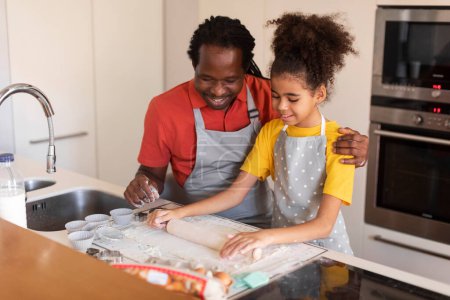 Foto de Feliz padre negro e hija rodando masa mientras hornean juntos en la cocina, sonriendo papá afroamericano y el niño femenino se divierten mientras preparan magdalenas en casa, disfrutando del tiempo juntos - Imagen libre de derechos