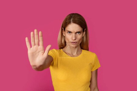 Photo for Serious sad confident adult caucasian woman blonde in yellow t-shirt shows palm hand, isolated on pink background, studio. People emotions, stop sign, gesture, against violence and covid-19 quarantine - Royalty Free Image