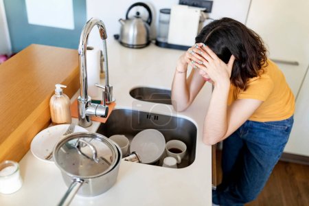 Foto de Mujer joven trastornada mirando un montón de platos sucios en el fregadero, mujer milenaria deprimida tocando la cabeza en la desesperación mientras limpia en la cocina en casa, cansado de hacer tareas domésticas, por encima de la vista - Imagen libre de derechos