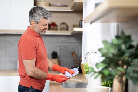 Photo for Positive handsome grey-haired middle aged man in casual outfits washing dishes at home, wearing colorful rubber gloves, side view, copy space. Household chores, house-keeping concept - Royalty Free Image