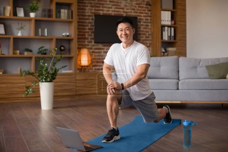 Foto de Emocionado hombre coreano maduro haciendo saltos, haciendo ejercicio para las piernas en el interior de la sala de estar, mirando a la cámara y sonriendo. Asiático masculino entrenamiento en frente de la computadora portátil, teniendo en línea lección con entrenador - Imagen libre de derechos