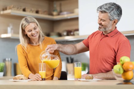 Photo for Handsome grey-haired mature man pouring fresh orange juice full of vitamin C for his smiling beautiful blonde wife, couple sitting at kitchen desk, having breakfast together, copy space - Royalty Free Image
