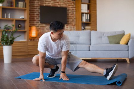 Téléchargez les photos : Entraînement à domicile. Coréen homme étirant les jambes sur tapis de yoga dans le salon, ayant l'entraînement domestique, espace libre. Homme actif faisant des exercices de flexibilité, garder la forme et en bonne santé, profiter de l'entraînement - en image libre de droit