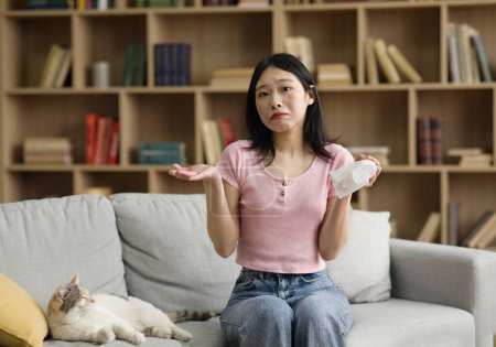 Téléchargez les photos : Femme coréenne bouleversée tenant des tissus en papier, allergique à la fourrure et souffrant d'écoulement nasal causé par son chat de compagnie, assise sur le canapé à la maison, espace de copie - en image libre de droit