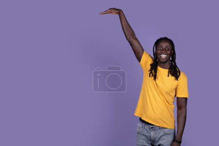 Téléchargez les photos : Joyeux beau jeune homme afro-américain aux cheveux longs en t-shirt jaune montrant la hauteur de quelque chose d'invisible avec sa main et souriant, posant sur fond de studio violet, espace de copie - en image libre de droit