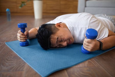 Téléchargez les photos : Fatigué asiatique mature homme couché sur tapis de fitness avec haltères dans les mains, se reposant après l'entraînement domestique dans le salon intérieur. Sport à la maison, concept de mode de vie actif - en image libre de droit