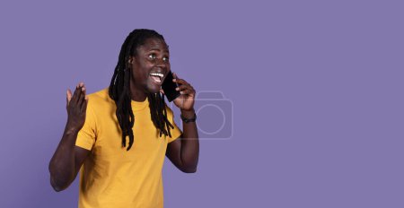 Téléchargez les photos : Émotionnel heureux mec noir élégant avec de longues dreadlocks en t-shirt jaune avoir une conversation téléphonique et gestuelle sur fond violet studio, partager des nouvelles passionnantes avec des amis, panorama, espace de copie - en image libre de droit
