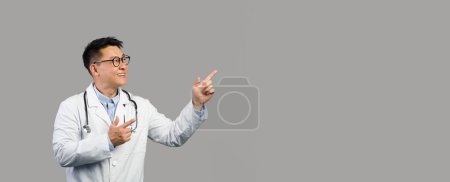 Téléchargez les photos : Joyeux homme chinois mature thérapeute en manteau blanc et lunettes pointant du doigt l'espace de copie, isolé sur fond gris, studio. Conseil médical, publicité et offre, traitement, soins de santé et médical - en image libre de droit