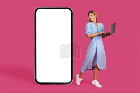 Photo for Smiling pretty adult caucasian blonde woman in dress with laptop looks at huge smartphone with blank screen, isolated on pink background, studio. Ad and offer, app for blog, study, work and business - Royalty Free Image