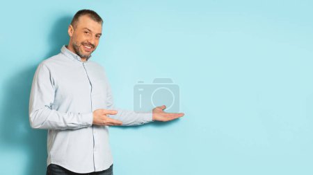 Photo for Happy handsome middle aged man in casual showing free space and advertising something on blue studio background, panorama with copy space - Royalty Free Image