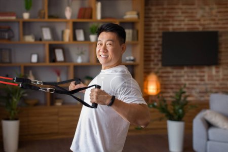 Téléchargez les photos : Actif asiatique d'âge moyen homme exerçant avec trx bandes de résistance à la maison, ayant l'entraînement domestique dans le salon intérieur, regardant et souriant à la caméra, espace libre - en image libre de droit