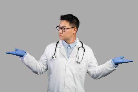 Photo for Serious mature chinese guy therapist in white coat and glasses spreads his arms to sides and holds empty space isolated on gray background. Doctor choice, ad and offer, health care during covid-19 - Royalty Free Image