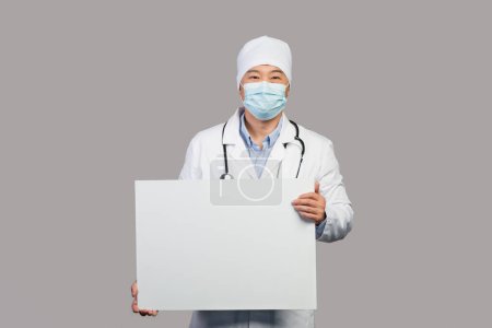 Photo for Smiling adult chinese male therapist in white coat with stethoscope, protective mask hold banner with copy space, isolated on gray background, studio. Doctor advice, medical examination, health care - Royalty Free Image