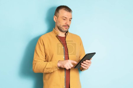 Photo for Modern technologies. Confident middle aged man holding and using digital tablet, standing over blue studio background. Serious mature male looking at gadget screen - Royalty Free Image