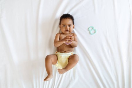 Téléchargez les photos : Petit bébé noir mignon portant une couche réutilisable jouant avec un jouet tout en étant allongé sur le lit, vue du dessus prise de vue d'un adorable bébé afro-américain ou d'une fille tenant une attache et regardant l'appareil photo, copiant l'espace - en image libre de droit