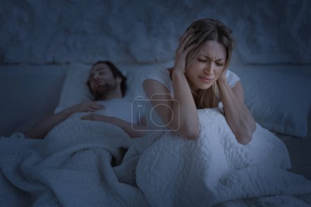 Photo for Depressed woman suffering from snoring sleeping husband, lady covering ears with hands, sitting on bed in the night. Noise, sleep problems, insomnia and apnea, copy space - Royalty Free Image