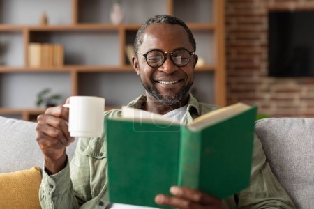 Photo for Happy mature black guy in glasses enjoys cup of coffee and reads book in free time in living room interior, close up, copy space. Hobby at weekend, literature for study and work, lifestyle at home - Royalty Free Image