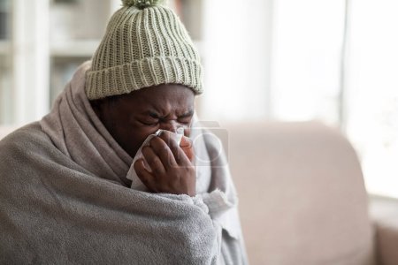 Téléchargez les photos : Gros plan de jeune homme noir malade couvert d'une couverture portant un chapeau tricoté assis sur le canapé dans le salon, éternuant son nez, utilisant des serviettes, souffrant de grippe saisonnière, espace de copie - en image libre de droit