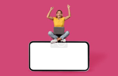 Photo for Cheerful excited surprised adult caucasian blonde lady with laptop raises hands, making victory, success gesture, sits on huge smartphone with blank screen, isolated on pink background. Ad and offer - Royalty Free Image