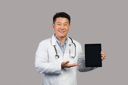 Photo for Happy mature chinese man therapist in white coat with stethoscope shows hand on tablet with empty screen, isolated on gray background, studio. Recommendation, medical support, health care and app - Royalty Free Image