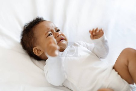 Photo pour Portrait de pleurer petit bébé noir portant une combinaison de corps allongé sur le lit à la maison, gros plan de bouleversé petit enfant afro-américain souffrant Colics ou dentition tout en se relaxant dans la chambre - image libre de droit