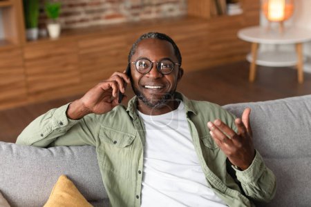 Téléchargez les photos : Heureux homme noir mature heureux dans les lunettes appelle par smartphone, parle, a de bonnes nouvelles à temps libre dans le salon intérieur, copier l'espace. Potins, communication à distance, émotions humaines et mode de vie à la maison - en image libre de droit