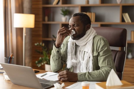 Photo for Unhappy sad adult black man with scarf blows his nose in napkin, suffers from runny nose and fever, works with computer in home office interior. Business and illness, flu and cold, feeling unwell - Royalty Free Image