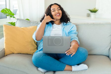 Téléchargez les photos : Heureuse femme noire plus la taille à l'aide d'un ordinateur portable et gestuelle appelez-moi, souriant à la caméra tout en travaillant et en communiquant en ligne, assis sur le canapé à la maison, espace libre - en image libre de droit