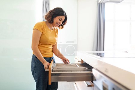 Téléchargez les photos : Jeune femme arabe rangeant les couverts dans le tiroir tout en nettoyant dans la cuisine à la maison, heureuse femme du Moyen-Orient assemblant des fourchettes, appréciant faire des tâches domestiques, vue latérale avec espace de copie - en image libre de droit