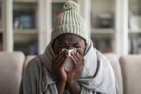 Photo for Closeup of ill young african american man covered in blanket wearing knitted ice hat sitting on sofa at home, sneezing his nose, using napkins, suffering from seasonal cold or flu, empty space - Royalty Free Image