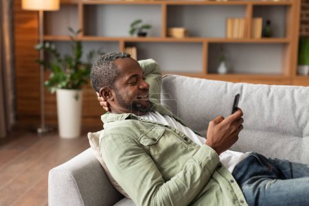 Photo for Glad mature black male uses smartphone, watch video has relax, surfing, read news, enjoy free time, lies on sofa in living room interior, copy space. New application, chat and social networks at home - Royalty Free Image