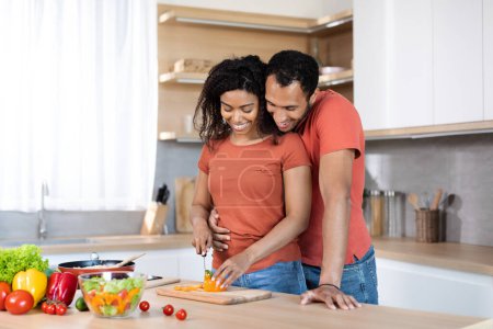 Foto de Feliz millennial hombre negro abrazando a la mujer en camiseta roja, dama ensalada de corte, preparando el almuerzo para la familia, cocinar juntos en el interior de la cocina. Relaciones, cuidado de la salud y comida casera en casa - Imagen libre de derechos