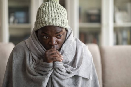 Foto de Primer plano del joven afroamericano frío cubierto de manta gris con sombrero de hielo de punto en su cabeza congelándose en casa, sentado en el sofá solo, calentando sus manos en la sala de estar, mirando el espacio de copia - Imagen libre de derechos
