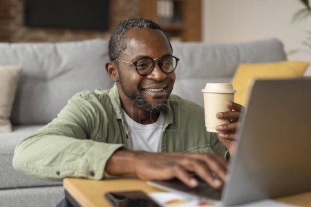 Foto de Trabajo y negocios en casa. Hombre negro adulto feliz en gafas de escribir en el ordenador, disfrutar de una taza de café para llevar en el interior de la sala de estar, de cerca. Tecnología moderna para la distancia social y la nueva normalidad - Imagen libre de derechos