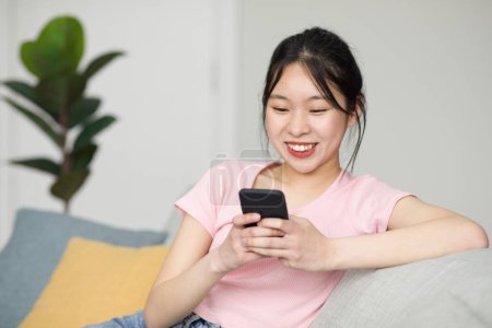 Téléchargez les photos : Femme coréenne excitée assise sur un canapé et utilisant un smartphone, bavardant avec des amis en ligne, espace de copie gratuit. Jolie femme avec téléphone portable refroidissant sur le canapé, vérifier les médias sociaux - en image libre de droit