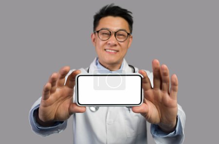 Foto de Glad hombre asiático de mediana edad terapeuta en bata blanca y gafas muestra teléfono inteligente con pantalla vacía, recomendar sitio web, aislado sobre fondo gris. Aplicación para la salud, el tratamiento de forma remota y nueva normal - Imagen libre de derechos
