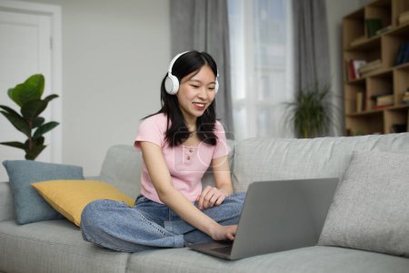 Téléchargez les photos : Jeune coréenne dans les écouteurs à l'aide d'un ordinateur portable, travaillant en ligne tout en étant assis sur le canapé à la maison, espace libre. Asiatique femelle ayant réunion à distance, parlant sur webcam - en image libre de droit