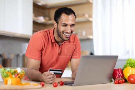 Photo for Glad handsome millennial black guy in red t-shirt uses credit card and computer for online shopping in kitchen interior. Ordering goods remotely, buy purchases, banking app, cashback and sale at home - Royalty Free Image