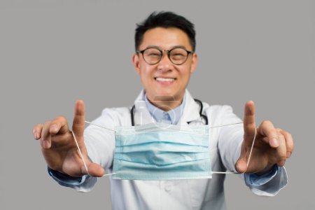 Photo for Happy middle aged asian male therapist in white coat and glasses puts mask on camera, isolated on gray background, studio. Health care advice, medical protection and support during covid-19 outbreak - Royalty Free Image