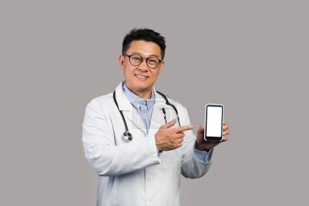 Photo for Smiling mature asian man in white coat and glasses points finger at smartphone with empty screen, isolated on gray background. Doctor advice and app recommendation, treatment remotely, new normal - Royalty Free Image