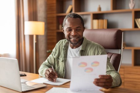 Photo for Cheerful busy middle aged black guy makes notes and works with graphs and charts, analysis financial data at workplace with computer in home office interior. Work and business with modern technology - Royalty Free Image