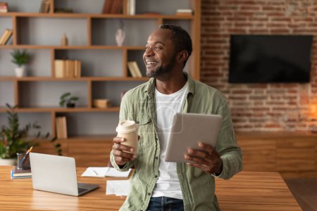 Téléchargez les photos : Homme noir adulte riant avec tablette et tasse de café à emporter montre à l'espace libre dans l'intérieur du bureau à la maison. Travail et affaires, analyse de données avec la technologie moderne, pause et repos avec une nouvelle application - en image libre de droit