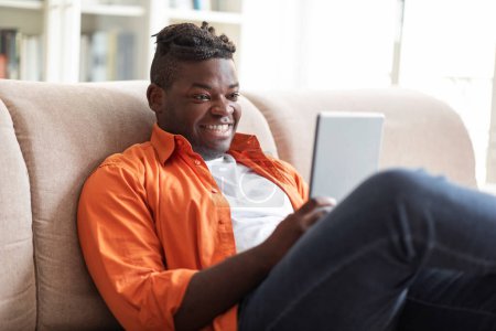 Téléchargez les photos : Portrait de heureux jeune homme potelé afro-américain avec une belle coiffure assise sur le canapé et utilisant une tablette numérique flambant neuve à la maison, regardant l'écran du pad et souriant, profitant d'une belle application, espace de copie - en image libre de droit