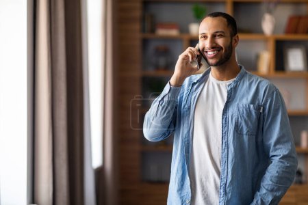 Foto de Retrato del guapo hombre negro sonriente hablando por teléfono celular en casa, alegre joven afroamericano parado cerca de la ventana en la sala de estar y disfrutando de una agradable conversación móvil, espacio para copiar - Imagen libre de derechos