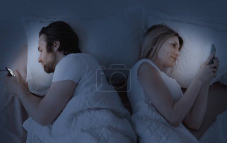 Foto de Concepto engañoso. Marido y esposa mensajes de texto en los teléfonos inteligentes, acostados espalda con espalda en la cama, ignorándose unos a otros, siendo indiferentes el uno al otro. Vista superior, poca luz - Imagen libre de derechos