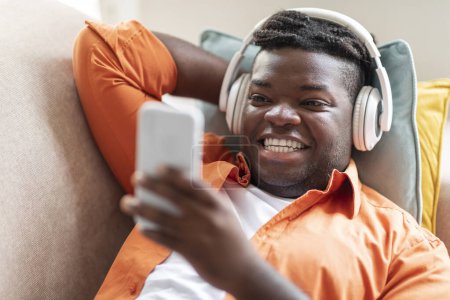 Photo for Happy Chubby African American Man With Nice Hairstyle Listening To Music On Mobile Phone Or Learning Via Online Podcast Wearing Headphones Relaxing On Sofa At Home. Mobile App For Music Lover Concept - Royalty Free Image