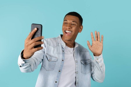 Photo for Cheerful excited young african american male with open mouth waving hand at webcam smartphone, has video call, say hello, hi, isolated on blue background, studio. Meeting remotely, chat and new app - Royalty Free Image